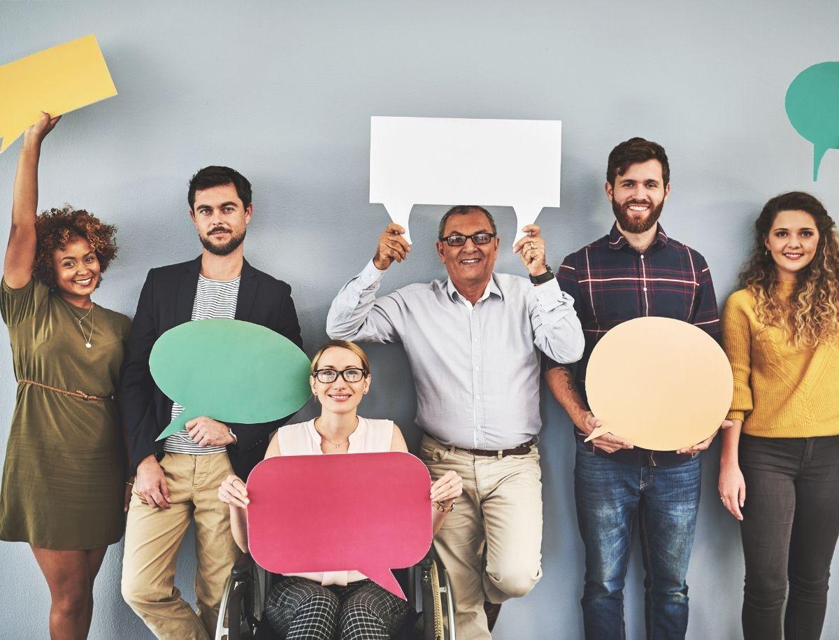 A diverse group of people stand against a gray wall, each holding a colorful, empty speech bubble cutout. One person is in a wheelchair.