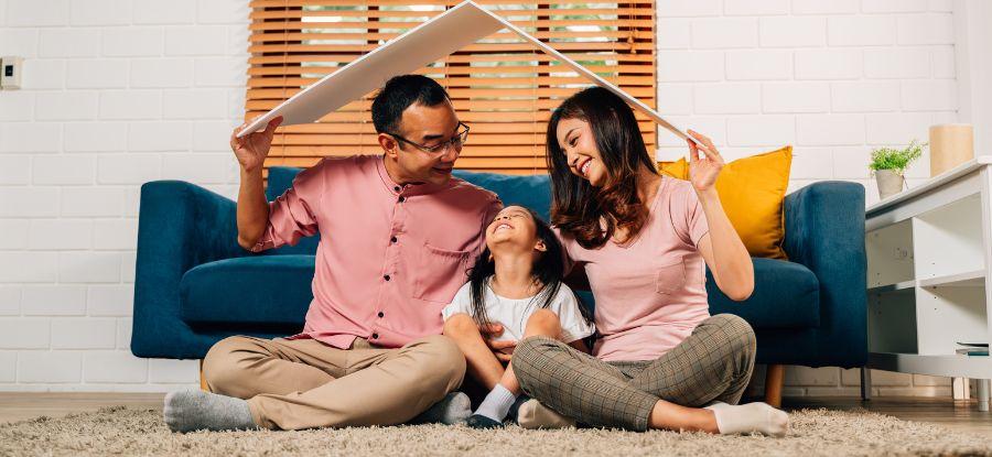 A family of three sits on the floor under a piece of cardboard held above them like a roof, symbolizing home protection.