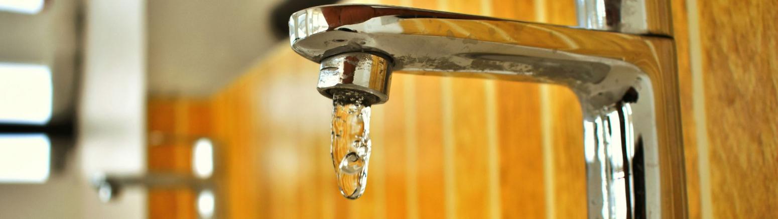 A close-up of a chrome faucet with water dripping from the spout against a blurred background of wooden paneling.