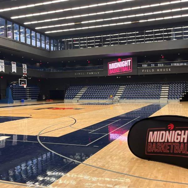 Angled view of a large indoor basketball court. A sign that says 'Midnight Basketball' is set up at the front corner of the court, and a light sign also reading 'Midnight Basketball' can be seen across the court above the stands. The court is empty, waiting for the program to begin.